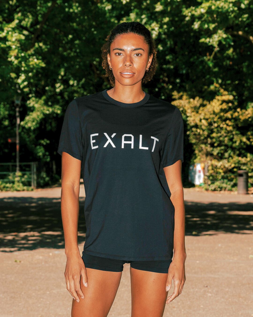 Close up shot of Women wearing Black EXALT x Lululemon Fundamental Tee in a park with trees in the background.