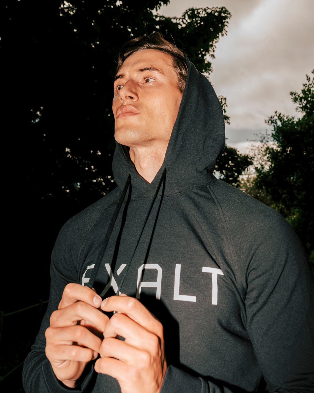 Close up shot of Man wearing Black EXALT x Lululemon City Sweat Hoodie with the hood up holding onto the drawstring. Background trees in a park.